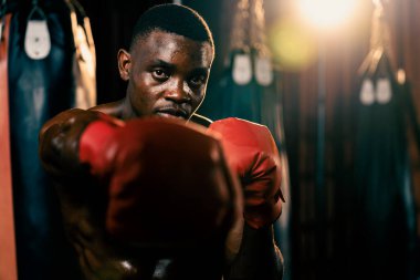 Üstsüz boksör pozu veren Afro-Amerikan Siyah Boksör yumruğunu agresif bir şekilde yumrukladı ve arka planda kum torbası ve boks ekipmanıyla spor salonunda dövüşmeye hazırdı. Impetus