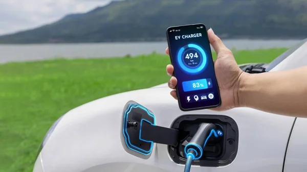 スマートフォンを持つビジネスマンは スマートEvモバイルアプリケーションによるバッテリーステータスインターフェースを表示し Ev車は自然湖の屋外の背景と充電ステーションから電気を充電します — ストック写真