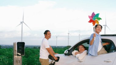 İlerici mutlu aile kavramı elektrikli araçla rüzgar çiftliğinde eğleniyor. Rüzgar türbini jeneratöründen gelen temiz yenilenebilir enerji ile şarj edilen elektrikli araç..