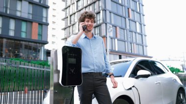 Takım elbiseli, hırslı bir işadamı elektrik kablosu ve yenilenebilir enerji enerjili bir elektrikli araçla şarj istasyonunda dururken elektrikli aracına yaslanıyor..