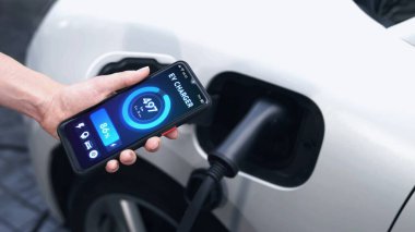 Akıllı telefon ekran bataryasını elinde tutan iş adamı akıllı EV mobil uygulama ile otoparktaki şarj istasyonundan elektrik şarj eden EV araba. İnceleyin
