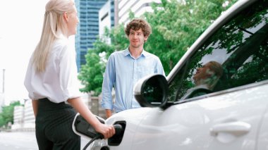 İlerici işadamı ve iş kadını, şehir merkezinde dolaşmadan önce şarj cihazını şarj istasyonundan elektrikli arabaya takıyor. Çevre dostu şarj edilebilir araba temiz enerjiyle çalışıyor..