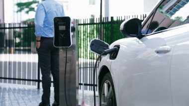 Takım elbiseli, hırslı bir işadamı elektrik kablosu ve yenilenebilir enerji enerjili bir elektrikli araçla şarj istasyonunda dururken elektrikli aracına yaslanıyor..