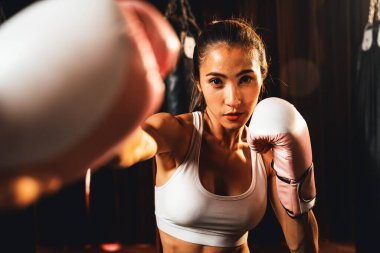 Asya 'lı kadın Muay Thai boksörü kamera önünde boksör yumruğu. Arka planda boks ekipmanıyla spor salonunda poz vermeye hazır. Kararlı gözler ve meydan okumaya hazır olun. Impetus