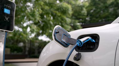 Elektrikli araba, çevre yeşil parkındaki elektrik kablosu ve yeşillik yeşili yeşillik yapraklarıyla şarj etmek için şarj istasyonuna bağlandı. Geleceğin yenilikçi her arabası ve enerji sürdürülebilirliği.