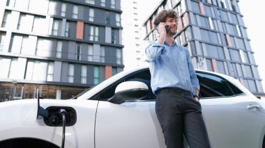 Takım elbiseli, hırslı bir işadamı elektrik kablosu ve yenilenebilir enerji enerjili bir elektrikli araçla şarj istasyonunda dururken elektrikli aracına yaslanıyor..