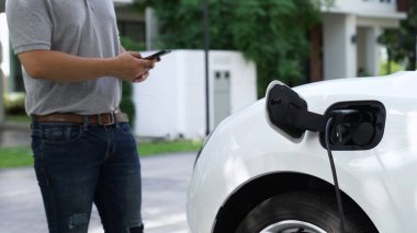 Elektrikli araba şarj istasyonuna bağlanıyor. Arka planda akıllı telefon kullanan adam EV şarj cihazıyla şarj oluyor. Sürdürülebilir temiz enerji kullanımının gelecekteki yaşam tarzı. İnceleyin