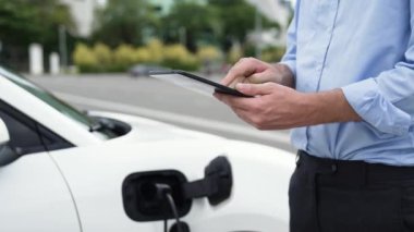 Modern iş adamı akıllı telefonun 3D hologramından batarya durumunu kontrol ederken EV arabasının şarj olan enerjisini kontrol ediyor. Gelecekteki EV otomotiv teknolojisinin yenilikçi teknolojik gelişimi. İnceleyin