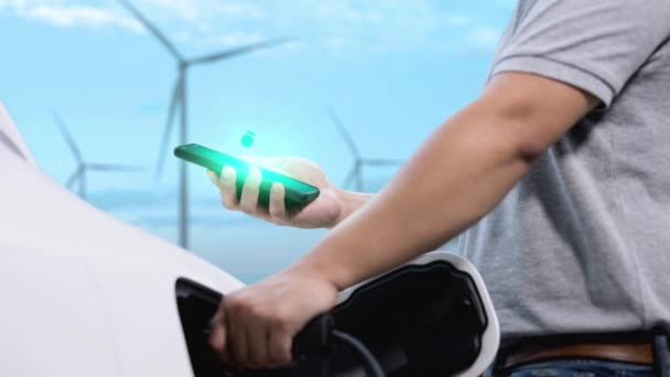 商人在智能手机上检查电池状态全息图显示 同时使用风力涡轮机发电机提供的替代清洁能源从充电站为其环保电动车充电 仔细阅读 — 图库视频影像