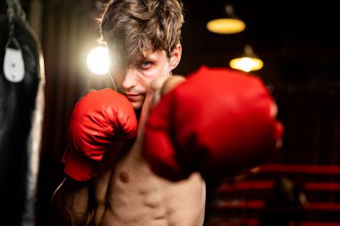 Üstsüz boksör pozu veren beyaz boksör, kameranın önünde agresif bir duruşta yumruğunu yumrukluyor ve arka planda kum torbası ve boks ekipmanıyla spor salonunda dövüşmeye hazır. Impetus