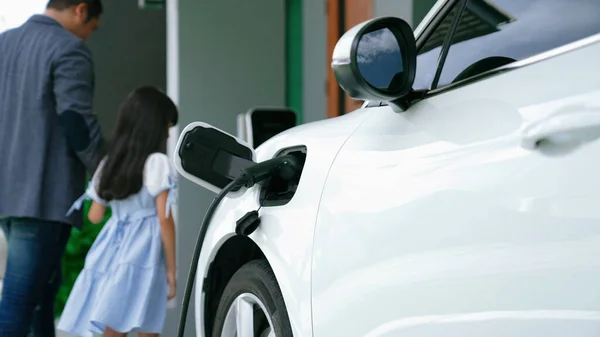 Progressive father and daughter installing plug from home charging station for electric vehicle. Future eco-friendly car with EV cars powered by renewable source of clean energy.