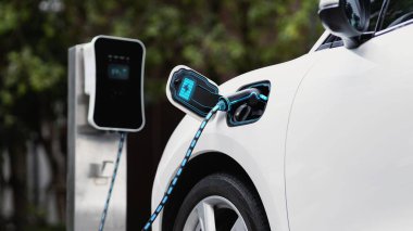 Elektrikli araba, çevre yeşil parkındaki elektrik kablosu ve yeşillik yeşili yeşillik yapraklarıyla şarj etmek için şarj istasyonuna bağlandı. Geleceğin yenilikçi her arabası ve enerji sürdürülebilirliği.