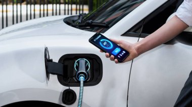 Akıllı telefonlu çalışan bir iş kadını akıllı EV mobil uygulama ile akü durumu arayüzünü tutarken EV arabası da otoparktaki şarj istasyonundan elektrik yüklüyor. İnceleyin