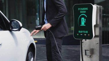 İş adamı elektrikli arabasını şehir merkezindeki şarj istasyonundan ya da otoparktan şarj ediyor. Şehir yaşam tarzında alternatif temiz enerji kullanan çevre dostu şarj edilebilir araba. İnceleyin.
