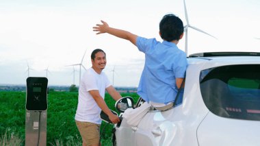 İlerici mutlu aile kavramı elektrikli araçla rüzgar çiftliğinde eğleniyor. Rüzgar türbini jeneratöründen gelen temiz yenilenebilir enerji ile şarj edilen elektrikli araç..