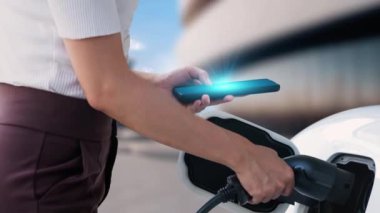 Modern bir kadın EV şarj cihazını EV 'nin şarj edildiği istasyondan şarj edilen arabasını şarj etmek için elektrikli otomobil portuna yerleştirir. Yenilikçi alternatif sürdürülebilir enerji kullanımı. İnceleyin