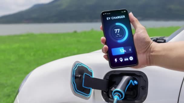 スマートフォンを持つビジネスマンは スマートEvモバイルアプリケーションによるバッテリーステータスインターフェースを表示し Ev車は自然湖の屋外の背景と充電ステーションから電気を充電します — ストック動画