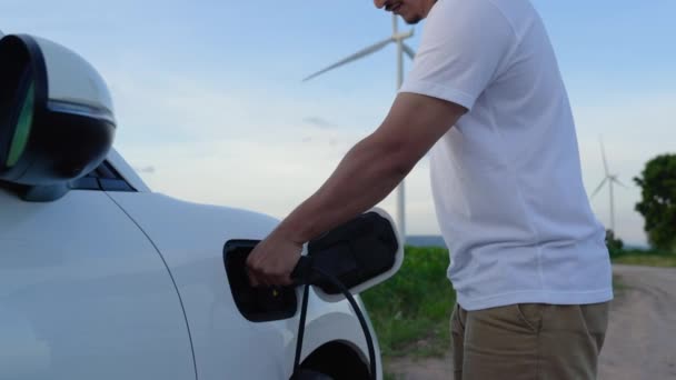 アジアの男は風力タービンによって動力を与えられる電気自動車にスマートEv充電器を挿入します 未来的なデジタルバッテリーステータスホログラムによる持続可能なクリーンEv技術のための再生可能エネルギー利用 パーセント — ストック動画