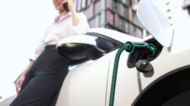 Akıllı telefon kullanan iş kadını, konut otoparkındaki şarj istasyonundan elektrikli arabayı şarj ediyor. Modern şehir yaşam tarzı ve karbondioksiti azaltmak için çevre dostu elektrikli araba kullanımı. İnceleyin