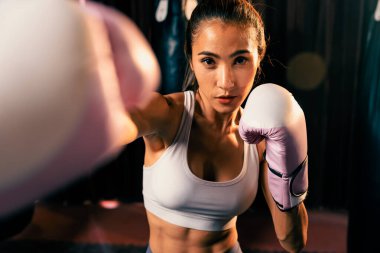 Asya 'lı kadın Muay Thai boksörü kamera önünde boksör yumruğu. Arka planda boks ekipmanıyla spor salonunda poz vermeye hazır. Kararlı gözler ve meydan okumaya hazır olun. Impetus