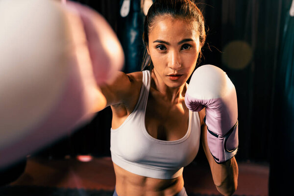 Asian female Muay Thai boxer punch fist in front of camera in ready to fight stance posing at gym with boxing equipment in background. Focused determination eyes and prepare for challenge. Impetus