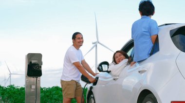 İlerici mutlu aile kavramı elektrikli araçla rüzgar çiftliğinde eğleniyor. Rüzgar türbini jeneratöründen gelen temiz yenilenebilir enerji ile şarj edilen elektrikli araç..