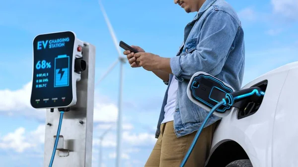 風力タービンファームの充電ステーションからEv充電器から電気自動車を充電しながらスマートフォンを使用するアジア人男性 持続可能でクリーンなエネルギー利用コンセプト パーセント — ストック写真