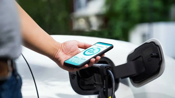 スマートフォンを持つビジネスマンはスマートEvモバイルアプリケーションによるバッテリーステータスインターフェースを表示し Ev車は駐車場の充電ステーションから電気を充電します Peruse — ストック写真