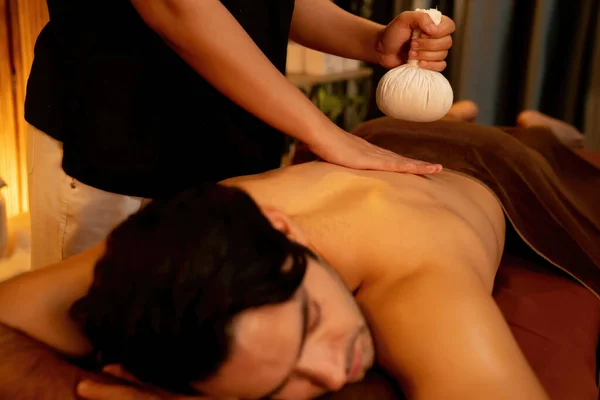 Hot herbal ball spa massage body treatment, masseur gently compresses herb bag on man body. Tranquil and serenity of aromatherapy recreation in warm lighting of candles at spa salon. Quiescent