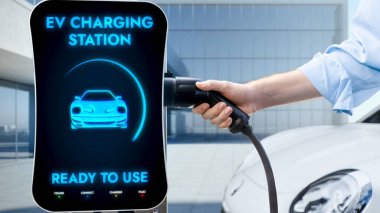 İşadamı, arka plandaki güneşli modern binadaki elektrikli araba şarj istasyonunu çek ve tut. Geleceğin temiz sürdürülebilir enerjisi ve EV araba teknolojik gelişimi.