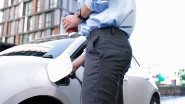 Takım elbiseli, hırslı bir işadamı elektrik kablosu ve yenilenebilir enerji enerjili bir elektrikli araçla şarj istasyonunda dururken elektrikli aracına yaslanıyor..