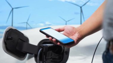 İş adamı akıllı telefon uygulamasında pil durumunu kontrol ederken rüzgar türbini jeneratöründen gelen alternatif temiz enerjiyi kullanarak çevre dostu EV 'yi şarj ediyor. İnceleyin
