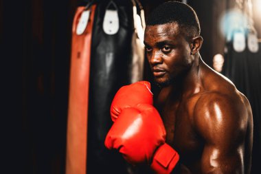 Boksör üstsüz, Afro-Amerikan siyah boksör savunma pozisyonu almış kırmızı eldiven giymiş, arka planda boks ekipmanları ve kum torbalarıyla spor salonunda dövüşmeye ve yumruk atmaya hazır. Impetus