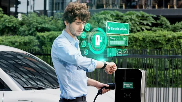 Businessman check EV car battery status on smartwatch hologram while recharge from charging station city car park. Futuristic lifestyle of clean energy for EV tech adaptation. Peruse