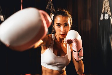 Asya 'lı kadın Muay Thai boksörü kamera önünde boksör yumruğu. Arka planda boks ekipmanıyla spor salonunda poz vermeye hazır. Kararlı gözler ve meydan okumaya hazır olun. Impetus