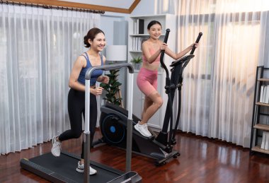 Enerjik ve güçlü atletik Asyalı kadın evde spor arkadaşı ya da antrenörü ile birlikte eliptik koşu makinesiyle koşuyor. Formda fiziğin takibi ve sağlıklı yaşam tarzına bağlılık. Kuvvetli