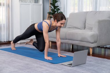 Spor kıyafetli Asyalı bir kadın antrenman rutini olarak spor minderinin üzerinde burpee yapıyor. Çekici kız sağlıklı yaşam tarzını online egzersiz videosuyla takip ediyor. Kuvvetli