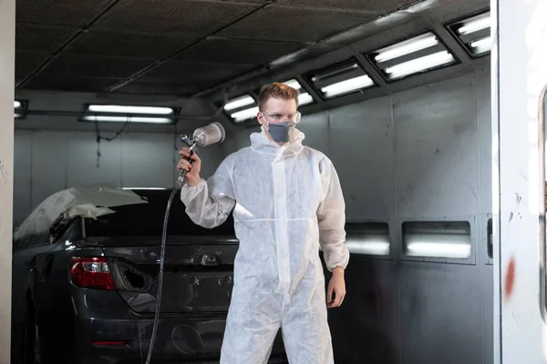 Automotive service worker in protective gear expertly applying color paint in to cars bodywork with spray gun or respirator painting in chamber workshop. Car paint service for scratch refinish. Oxus