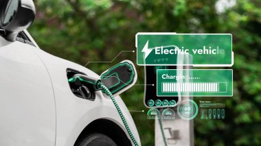 EV şarj kablosuna elektrik kablosu takılı. Çevre yeşil parkı ve yeşillik arka planında akıllı dijital batarya durum hologramını gösteriyor. Enerji sürdürülebilirliği teknolojik gelişim.
