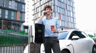 Takım elbiseli, hırslı bir işadamı elektrik kablosu ve yenilenebilir enerji enerjili bir elektrikli araçla şarj istasyonunda dururken elektrikli aracına yaslanıyor..