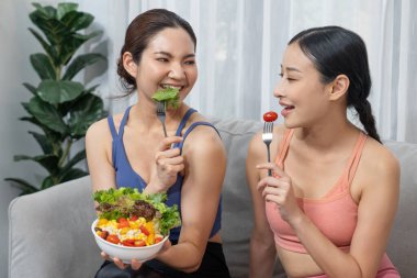 Spor kıyafetleri içinde, ellerinde salata kasesi olan genç Asyalı kadınlar canlı meyve ve sebzelerle dolular. Doğal gençlik ve yaşam tarzı dengeli beslenme ve ev egzersizi ile uyumlu. Kuvvetli