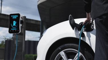 İş kadını, şehir merkezindeki şarj istasyonundan ya da kamu otoparkından elektrikli arabasını şarj ediyor. Alternatif temiz enerji kullanan çevre dostu şarj edilebilir araba. İnceleyin