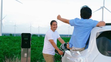 İlerici mutlu aile kavramı elektrikli araçla rüzgar çiftliğinde eğleniyor. Rüzgar türbini jeneratöründen gelen temiz yenilenebilir enerji ile şarj edilen elektrikli araç..