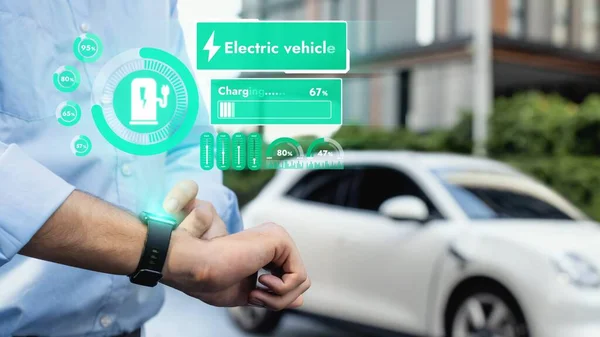 Businessman checking EV car battery status in hologram from smartwactch while recharging from charging station. Modern urban lifestyle and eco-friendly electric car utilization to reduce CO2. Peruse