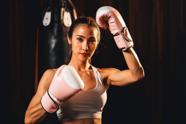 Asya 'lı kadın Muay Thai boksörü kamera önünde boksör yumruğu. Arka planda boks ekipmanıyla spor salonunda poz vermeye hazır. Kararlı gözler ve meydan okumaya hazır olun. Impetus