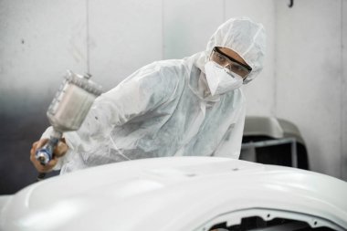 Tam korumalı ekipmanlı otomotiv işçisi sprey tabanca ya da oda atölyesindeki solunum cihazı boyasıyla araba kaportasına ustalıkla renk boyası uygular. Tamir için araba boyama servisi. Oxus.