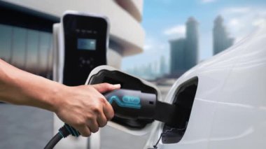 Adam elektrikli arabaya akıllı EV şarj cihazı takıyor ve arka planda modern şehir manzarası olan EV şarj istasyonundan şarj oluyor. ESG şehrinde gelecekteki sürdürülebilir güç ve EV araba kullanımı. İnceleyin