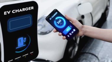 Akıllı telefon akü durumu arayüzünü elinde tutan iş kadını akıllı EV mobil uygulama ile otomobil parkındaki şarj istasyonundan elektrik şarj eden çevre dostu EV araba. İnceleyin