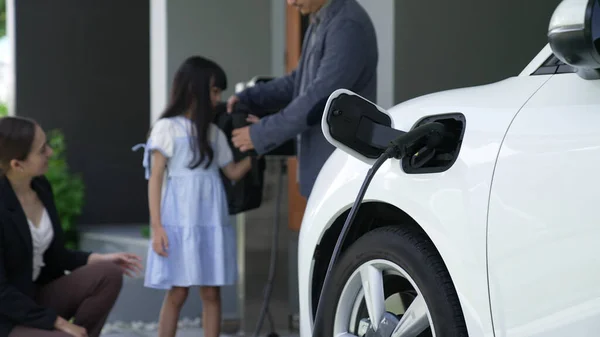 Progressive parents with electric vehicle and home charging station. Happy family with daughter giving each other high fives before leave for school. Alternative future transportation concept of EVs