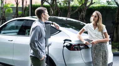 Genç çift yazın yeşil sürdürülebilir şehir bahçesinde EV elektrikli araba ile seyahat ederken, yeşil temiz yeniden şarj edilebilir elektrikli araç iç organları ile kentsel sürdürülebilirlik yaşam tarzını gösteriyor.